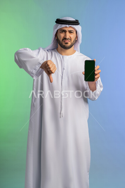 Thumbs down gesture of rejection and disapproval, concept of negative evaluation and annoyance, empty green screen chroma key display via mobile, portrait of an Arab Gulf Emirati man wearing a white kandura and ghutra holding a mobile phone, using a modern and advanced technical device, colorful background