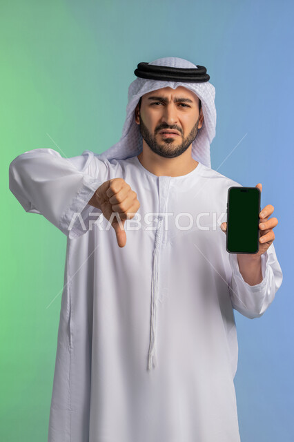 الاشارة باصبع الابهام للأسفل بايماءات الرفض وعدم القبول، مفهوم التقييم السلبي والانزعاج، عرض شاشة خضراء فارغة كروما عبر الجوال، بورتريه لرجل عربي خليجي اماراتي يرتدي الكندورة والغترة البيضاء يمسك بيده الهاتف المحمول، استخدام جهاز تقني حديث ومتطور، خلفية ملونة