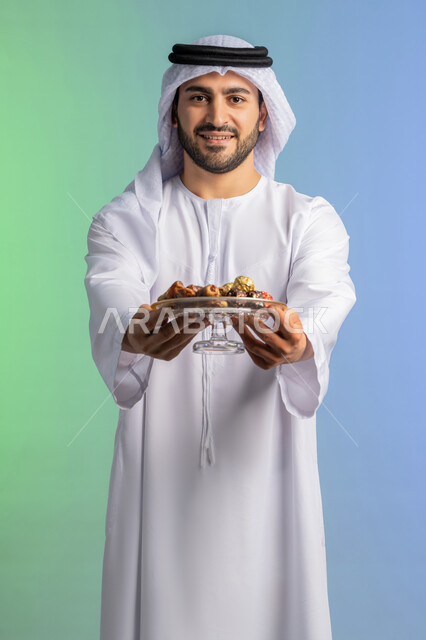 افطار الصائم في رمضان، ضيافة رمضانية لذيذة، منتج ومحصول محلي وطني، وجبة ذات قيمة غذائية عالية، بورتريه مقرب لرجل عربية خليجية اماراتي يرتدي الكندورة والغترة يحمل بيده طبق من التمر الصحي وينظر الى الكاميرا، الاحتفال بقدوم شهر الخير