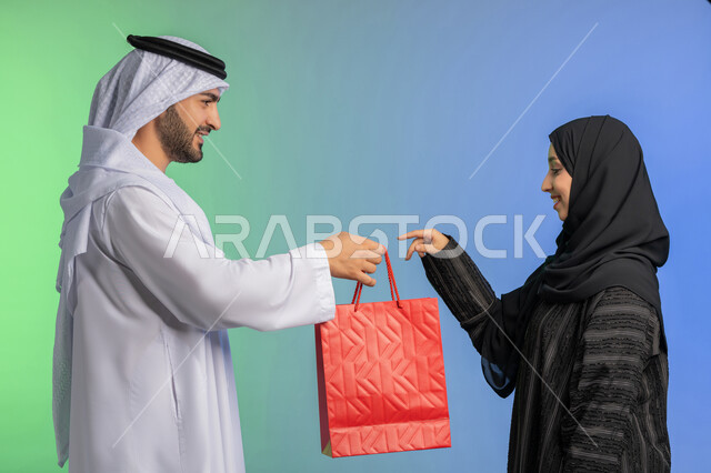 استلام طلبات اونلاين، مفهوم التسوق واقتناء الهدايا، الاستفادة من العروض في شراء الحاجات الضرورية، بورتريه  لرجل عربي خليجي اماراتي  يعطي زوجته كيس ورقي، خلفية ملونة