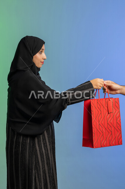 الاستفادة من العروض في شراء الحاجيات الضرورية، مفهوم التسوق واقتناء الهدايا، استلام طلبات اونلاين، بورتريه من الجانب لمرأة عربية خليجية إماراتيه ترتدي العباءة والحجاب تمسك بيدها كيس ورقي، خلفية ملونة