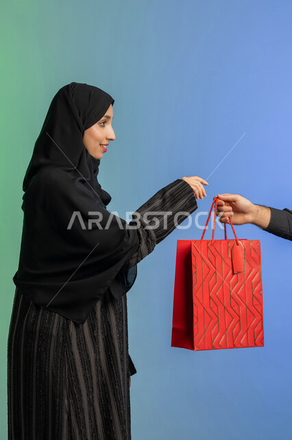 مفهوم التسوق واقتناء الهدايا، الاستفادة من العروض في شراء الحاجات الضرورية، استلام طلبات اونلاين، بورتريه من الجانب لمرأة عربية خليجية إماراتيه ترتدي العباءة والحجاب تمسك بيدها كيس ورقي، خلفية ملونة