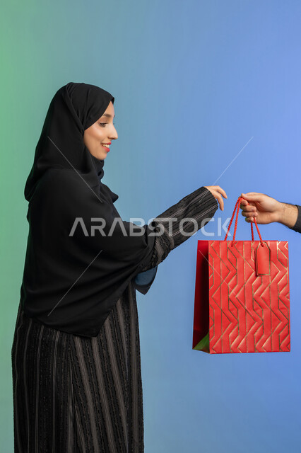الاستفادة من العروض في شراء الحاجيات الضرورية، مفهوم التسوق واقتناء الهدايا، استلام طلبات اونلاين، بورتريه من الجانب لمرأة عربية خليجية إماراتيه ترتدي العباءة والحجاب تمسك بيدها كيس ورقي، خلفية ملونة