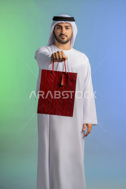 شراء الحاجات والهدايا، الاستفادة من العروضات والتخفيضات، متعة التسوق في المولات التجارية، النظر إلى الكاميرا بإيماءات البهجة والسرور، بورتريه لرجل عربي خليجي إماراتي مبتسم يرتدي الكندورة والغترة يحمل بيديه كيس ورقي، خلفية ملونة
