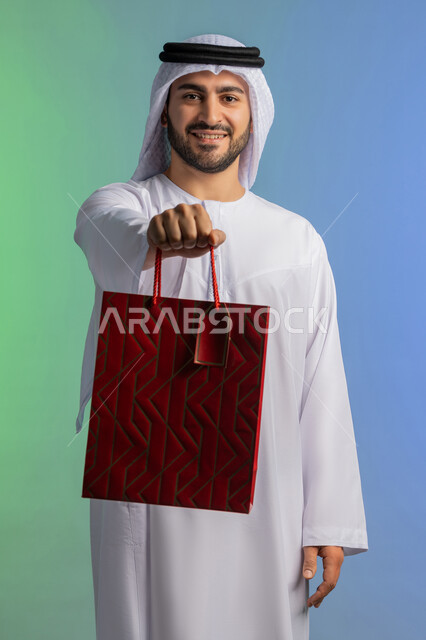 متعة التسوق في المولات التجارية، شراء الحاجات والهدايا، الاستفادة من العروضات والتخفيضات، النظر إلى الكاميرا بإيماءات البهجة والسرور، بورتريه مقرب لرجل عربي خليجي إماراتي مبتسم يرتدي الكندورة والغترة يحمل بيديه كيس ورقي، خلفية ملونة