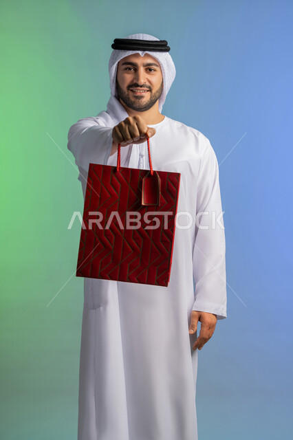 شراء الحاجات والهدايا، الاستفادة من العروضات والتخفيضات، متعة التسوق في المولات التجارية، النظر إلى الكاميرا بإيماءات البهجة والسرور، بورتريه لرجل عربي خليجي إماراتي مبتسم يرتدي الكندورة والغترة يحمل بيديه كيس ورقي، خلفية ملونة