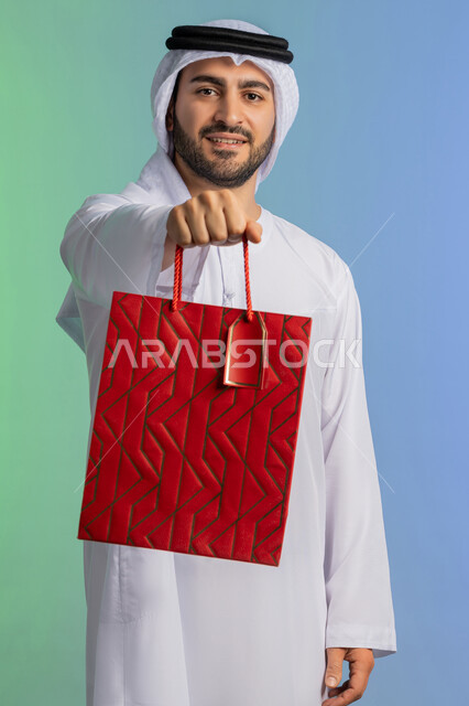 متعة التسوق في المولات التجارية، شراء الحاجات والهدايا، الاستفادة من العروضات والتخفيضات، النظر إلى الكاميرا بإيماءات البهجة والسرور، بورتريه مقرب لرجل عربي خليجي إماراتي مبتسم يرتدي الكندورة والغترة يحمل بيديه كيس ورقي، خلفية ملونة