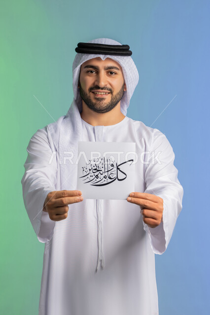 Preparing to welcome the holy month of Ramadan, the joy and happiness of Muslims with the arrival of the month of goodness, a portrait of a smiling Emirati Gulf Arab man wearing a kandura and a ghutra displaying a manuscript of “Happy New Year” decorated with gestures of joy and happiness, a colorful background