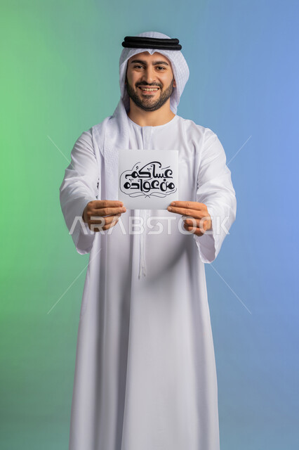 بهجة وفرحة المسلمين بقدوم شهر الخير، بورتريه لرجل عربي خليجي اماراتي مبتسم يرتدي الكندورة والغترة  يقوم بعرض مخطوطة عساكم من عواده مزخرفة بإيماءات السرور والسعادة, الاستعداد لاستقبال شهر رمضان المبارك, خلفية ملونة