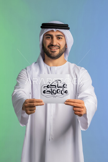 الاستعداد لاستقبال شهر رمضان المبارك, بهجة وفرحة المسلمين بقدوم شهر الخير، بورتريه لرجل عربي خليجي اماراتي مبتسم يرتدي الكندورة والغترة يقوم بعرض مخطوطة عساكم من عواده مزخرفة بإيماءات السرور والسعادة,  خلفية ملونة