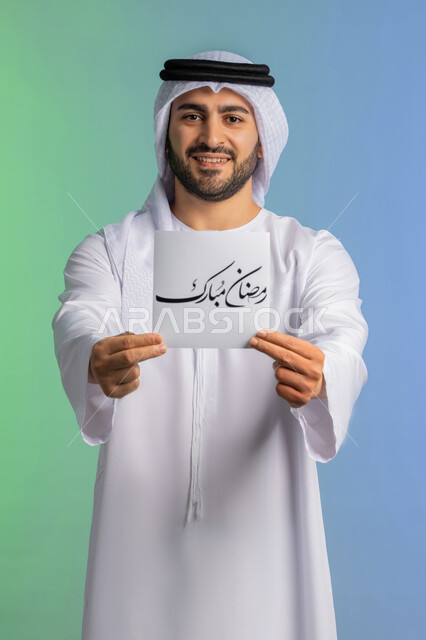 بهجة وفرحة المسلمين بقدوم شهر الخير، بورتريه لرجل عربي خليجي اماراتي مبتسم يرتدي الكندورة والغترة  يقوم بعرض مخطوطة رمضان مبارك مزخرفة بإيماءات السرور والسعادة, الاستعداد لاستقبال شهر رمضان المبارك, خلفية ملونة