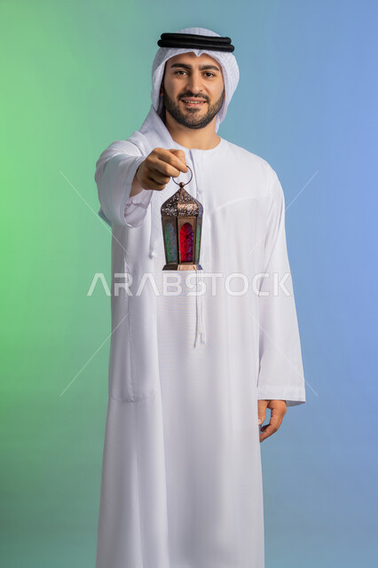 استقبال شهر الخير والبركة, المناسبات الدينية والأعياد الإسلامية, الاستعداد لقدوم شهر رمضان, بورتريه لرجل عربي خليجي اماراتي يرتدي الغترة والكندورة التقليدية يحمل الفانوس وينظر الى الكاميرا بتعابير الفرح, تحف وزينة الاحتفالات في السعودية, خلفية ملونة