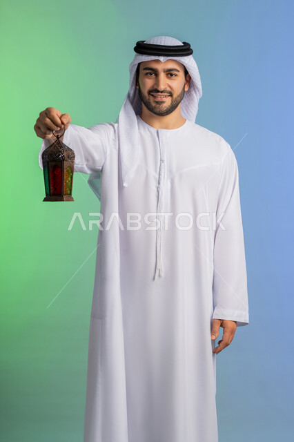 Religious occasions and Islamic holidays, welcoming the month of goodness and blessings, preparing for the arrival of the month of Ramadan, portrait of an Arab Gulf Emirati man wearing the traditional ghutra and kandura, holding a lantern and looking at the camera with expressions of joy, festive decorations and artifacts in Saudi Arabia, colorful background