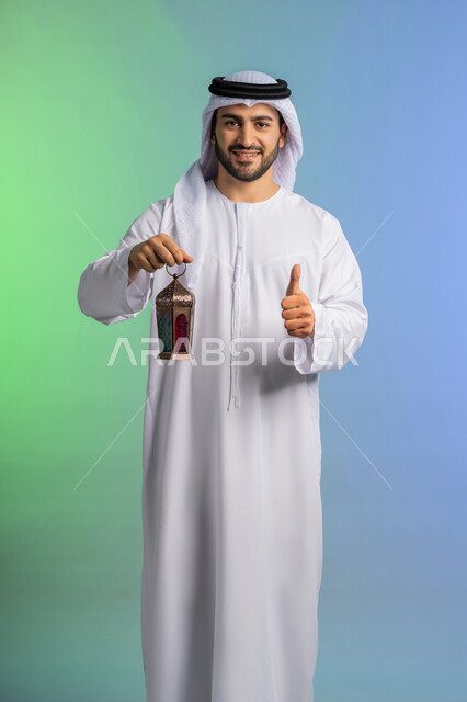 استقبال شهر الخير والبركة, تعابير باصبع الابهام تدل على الاعجاب والقبول، المناسبات الدينية والأعياد الإسلامية, الاستعداد لقدوم شهر رمضان, بورتريه لرجل عربي خليجي اماراتي يرتدي الغترة والكندورة التقليدية يحمل الفانوس وينظر الى الكاميرا بتعابير الفرح, تحف وزينة الاحتفالات في السعودية, خلفية ملونة