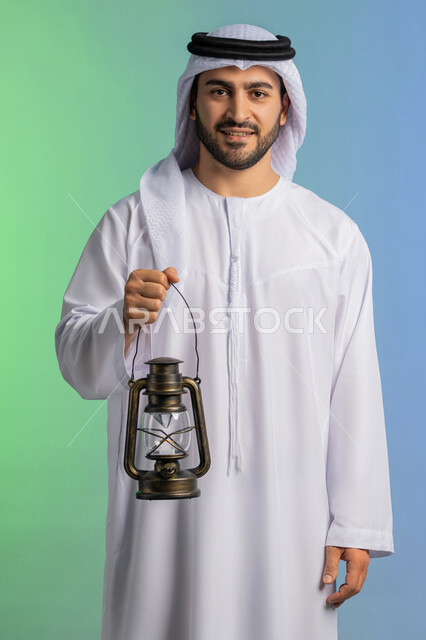 Welcoming the month of goodness and blessings, religious occasions and Islamic holidays, preparing for the arrival of the month of Ramadan, portrait of an Arab Gulf Emirati man wearing the traditional ghutra and kandura, holding a lantern and looking at the camera with expressions of joy, festive decorations and artifacts in Saudi Arabia, colorful background