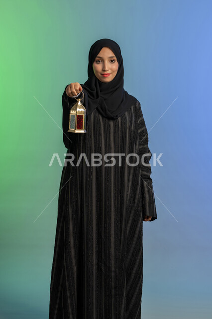 الأعياد والمناسبات الدينية، أجواء السكينة والروحانية، بورتريه لسيدة عربية خليجية اماراتية محجبة ترتدي العباءة السوادء والحجاب تحمل بيدها فانوس معدني تنظر الى الكاميرا بايماءات السعادة والسرور، الإستعداد لقدوم شهر رمضان المبارك، العبادة والتقرب إلى الله، خلفية ملونة