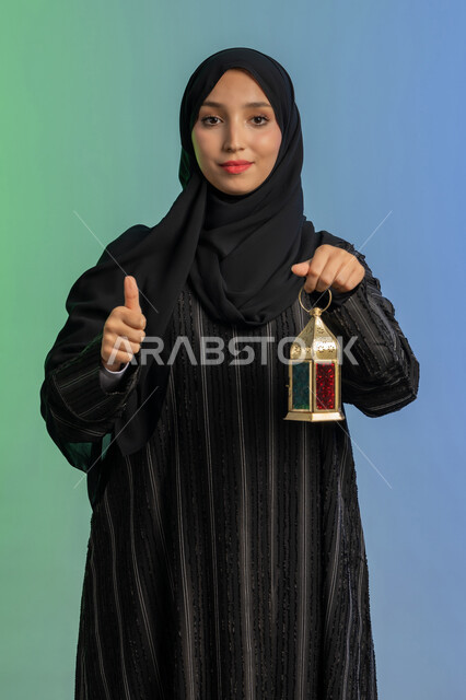 الأعياد والمناسبات الدينية، أجواء السكينة والروحانية، بورتريه لسيدة عربية خليجية اماراتية محجبة ترتدي العباءة السوادء والحجاب تحمل بيدها فانوس معدني تنظر الى الكاميرا بايماءات السعادة والسرور، الإستعداد لقدوم شهر رمضان المبارك، العبادة والتقرب إلى الله، خلفية ملونة
