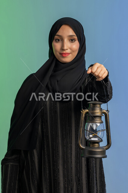 أجواء السكينة والروحانية،  الاحتفال بالأعياد والمناسبات الدينية، بورتريه لسيدة عربية خليجية اماراتية محجبة ترتدي العباءة السوادء والحجاب تحمل بيدها فانوس معدني تنظر الى الكاميرا بايماءات السعادة والسرور، الإستعداد لقدوم شهر رمضان المبارك، العبادة والتقرب إلى الله، خلفية ملونة