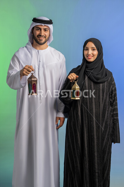 المناسبات الدينية والأعياد الإسلامية, بورتريه لرجل عربي خليجي اماراتي يرتدي الكندورة ويضع الغترة يقف مع زوجته ويحملان بايديهم فانوس رمضاني, سيدة عربية خليجية اماراتية ترتدي العباءة السوداء والحجاب تحتفل بقدوم الشهر الفضيل, الإستعداد لرمضان المبارك, خلفية ملونة
