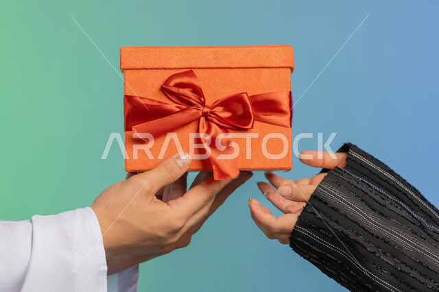 The concept of offering gifts on holidays and occasions, the husband presents his wife with a gift on the occasion of Valentine’s Day, a portrait of an Arab Gulf Emirati man wearing a kandora and a ghutra offering the gift, an Arab Gulf Emirati woman wearing a black abaya and hijab standing with facial and hand gestures indicating astonishment and joy, a colorful background