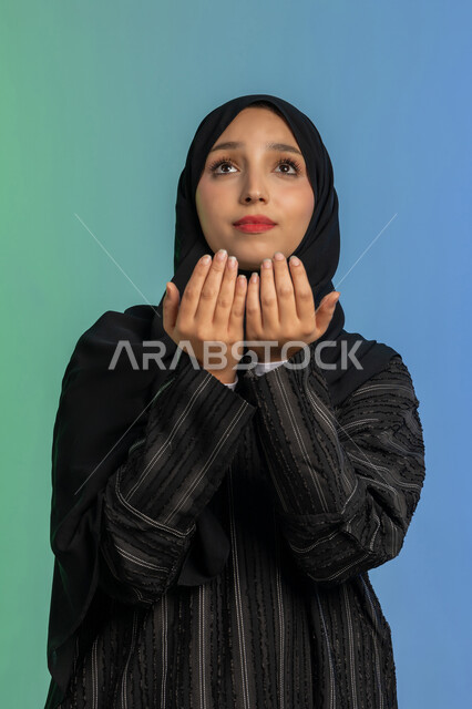التمني والطلب من الله، ايماءات تدل على الخشوع والتضرع، الاجتهاد في الطاعات والأدعية والأذكار، العبادة والتقرب الى الله، بورتريه مقرب لإمرأة عربية خليجية اماراتية ترتدي الحجاب والعباءة ترفع يديها وتقوم بالدعاء، خلفية ملونة