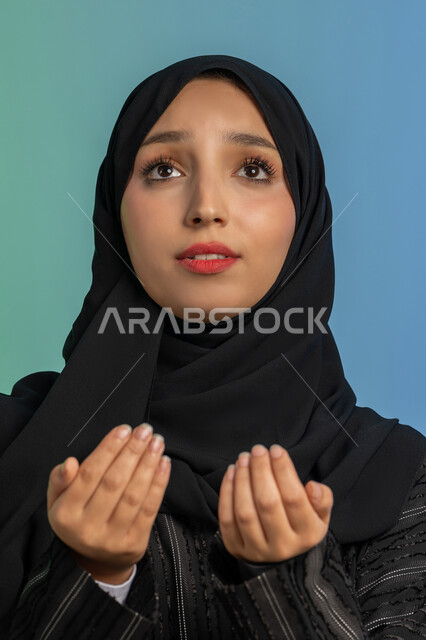 ايماءات تدل على الخشوع والتضرع، التمني والطلب من الله، الاجتهاد في الطاعات والأدعية والأذكار، العبادة والتقرب الى الله، بورتريه مقرب لإمرأة عربية خليجية اماراتية ترتدي الحجاب والعباءة ترفع يديها وتقوم بالدعاء، خلفية ملونة