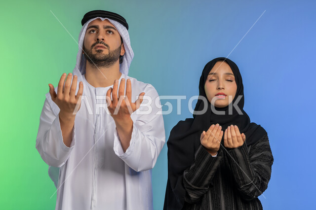 التمني والطلب من الله، ايماءات تدل على الخشوع والتضرع، الاجتهاد في الطاعات والأدعية والأذكار، العبادة والتقرب الى الله، بورتريه مقرب لزوجان  عربيان خليجيان اماراتيان يقومان برفع ايديهم بايماءات الدعاء، خلفية ملونة