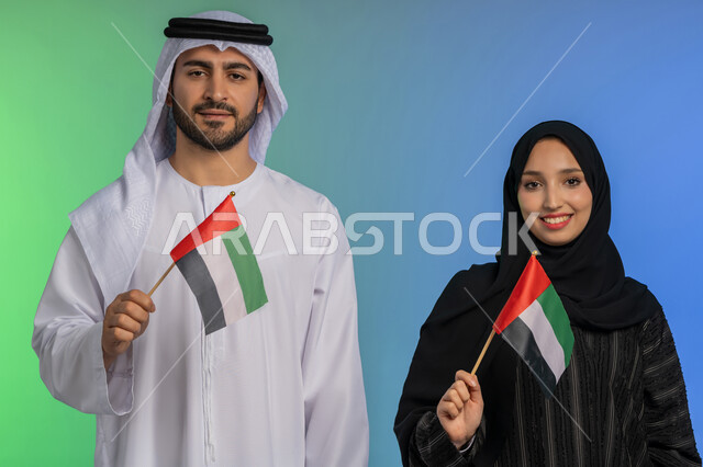 مفهوم حب الوطن والعلم، الاحتفال باليوم الوطني الإماراتي 2 ديسمبر، إحياء ذكرى استقلال دولة الإمارات العربية المتحدة، بورتريه لرجل وامراة عربيان خليجيان اماراتيان يحملان علم الإمارات، الاعتزاز بإنجازات الوطن، النظر الى الكاميرا بايماءات السعادة والفخر،  خلفية ملونة