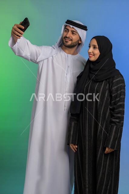 Emirati man wearing kandura and ghutra raising his hand with the number three gesture, expressions of happiness and pleasure, documenting happy moments via mobile, portrait of a smiling veiled Emirati Gulf Arab woman wearing a black abaya taking a commemorative selfie with her husband, colorful background