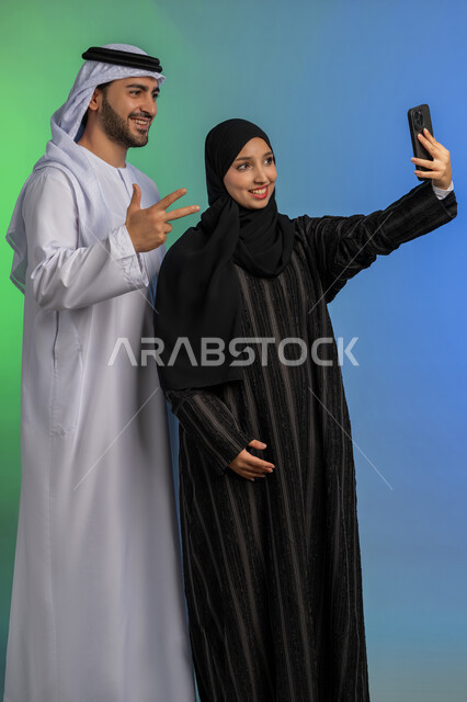رجل اماراتي يرتدي الكندورة والغترة يرفع يده بايماءات الرقم ثلاثة، تعابير السعادة والسرور, توثيق اللحظات السعيدة عبر الجوال، بورتريه لامراة عربية خليجية اماراتية محجبة مبتسمة ترتدي العباءة السوداء تقوم بالتقاط صورة سيلفي تذكارية مع زوجها، خلفية ملونة