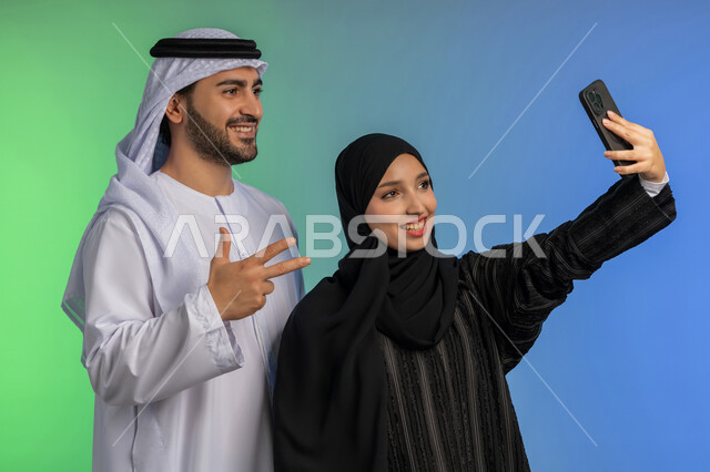 تعابير السعادة والسرور, توثيق اللحظات السعيدة عبر الجوال، بورتريه مقرب لامراة عربية خليجية اماراتية محجبة مبتسمة ترتدي العباءة السوداء تقوم بالتقاط صورة سيلفي تذكارية مع زوجها، رجل اماراتي يرتدي الكندورة والغترة يرفع يده بايماءات الرقم ثلاثة، خلفية ملونة