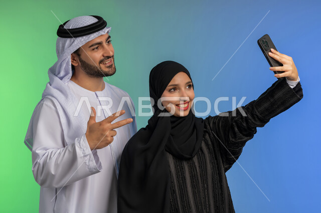 توثيق اللحظات السعيدة عبر الجوال، بورتريه مقرب لامراة عربية خليجية اماراتية محجبة مبتسمة ترتدي العباءة السوداء تقوم بالتقاط صورة سيلفي تذكارية مع زوجها، رجل اماراتي يرتدي الكندورة والغترة يرفع يده بايماءات الرقم ثلاثة، تعابير السعادة والسرور, خلفية ملونة