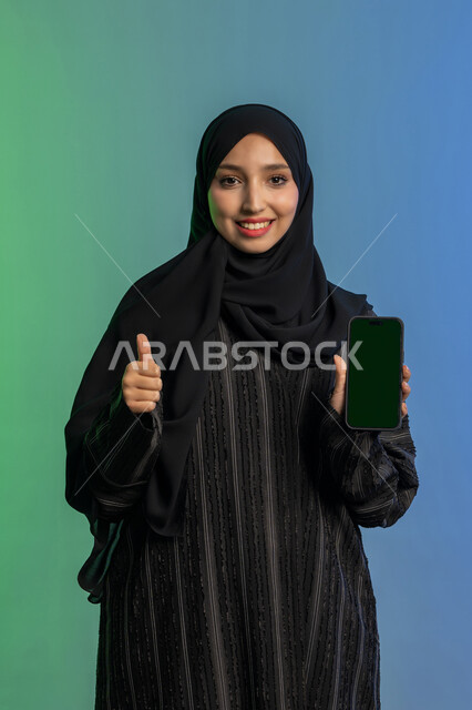 مفهوم القبول والإعجاب والرضا, رفع اصبع الابهام للأعلى بايماءات الجودة والامتياز، عرض تطبيقات الجوال عبر شاشة خضراء فارغة كروما، بورتريه لامرأة عربية خليجية اماراتية محجبة ترتدي العباءة السوداء تمسك بيدها الهاتف المحمول، استخدام جهاز تقني حديث ومتطور، خلفية ملونة