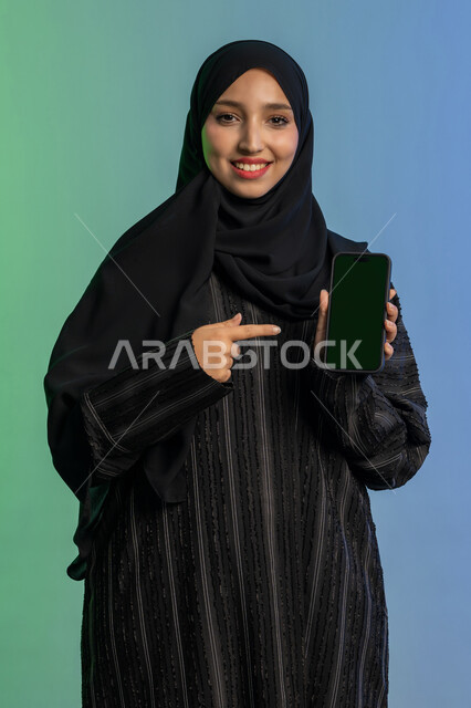 النظر الى الكاميرا بايماءات السعادة والسرور، عرض تطبيقات الجوال، الاشارة باصبع السبابة الى شاشة خضراء فارغة كروما،  بورتريه لامرأة عربية خليجية اماراتية محجبة ترتدي العباءة السوداء تمسك بيدها الهاتف المحمول، استخدام جهاز تقني حديث ومتطور، خلفية ملونة