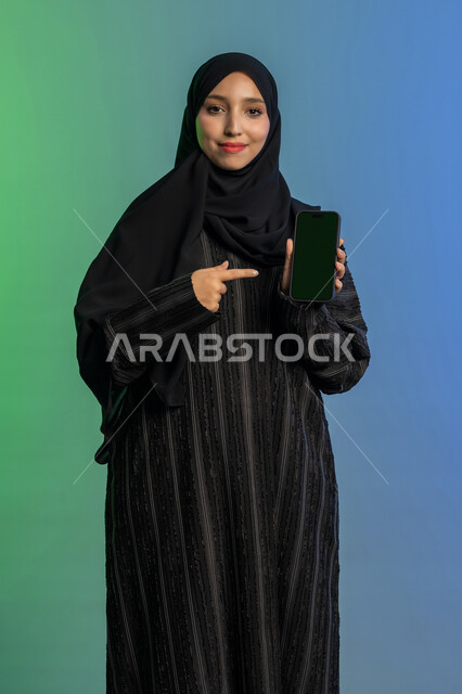 الاشارة باصبع السبابة الى شاشة خضراء فارغة كروما،  استخدام جهاز تقني حديث ومتطور، النظر الى الكاميرا بايماءات السعادة والسرور، عرض تطبيقات الجوال، بورتريه لامرأة عربية خليجية اماراتية محجبة ترتدي العباءة السوداء تمسك بيدها الهاتف المحمول، خلفية ملونة