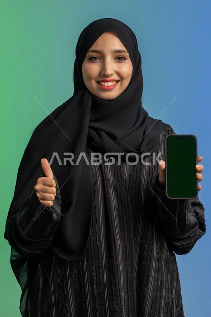مفهوم القبول والإعجاب والرضا, رفع اصبع الابهام للأعلى بايماءات الجودة والامتياز، عرض تطبيقات الجوال عبر شاشة خضراء فارغة كروما، بورتريه لامرأة عربية خليجية اماراتية محجبة ترتدي العباءة السوداء تمسك بيدها الهاتف المحمول، استخدام جهاز تقني حديث ومتطور، خلفية ملونة