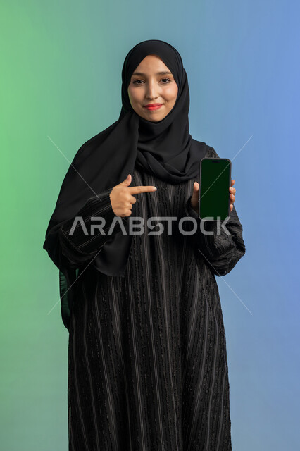 النظر الى الكاميرا بايماءات السعادة والسرور، عرض تطبيقات الجوال، الاشارة باصبع السبابة الى شاشة خضراء فارغة كروما،  بورتريه لامرأة عربية خليجية اماراتية محجبة ترتدي العباءة السوداء تمسك بيدها الهاتف المحمول، استخدام جهاز تقني حديث ومتطور، خلفية ملونة