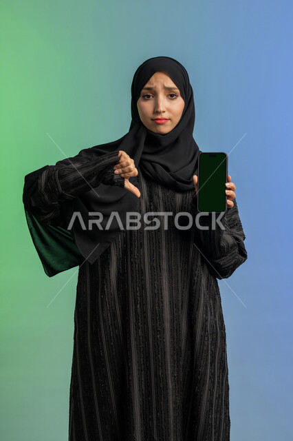 Thumbs down gesture of rejection and disapproval, concept of negative evaluation and annoyance, empty green screen display chroma key on mobile, portrait of veiled Arab Gulf Emirati woman wearing black abaya holding mobile phone in hand, using modern and advanced technical device, colorful background