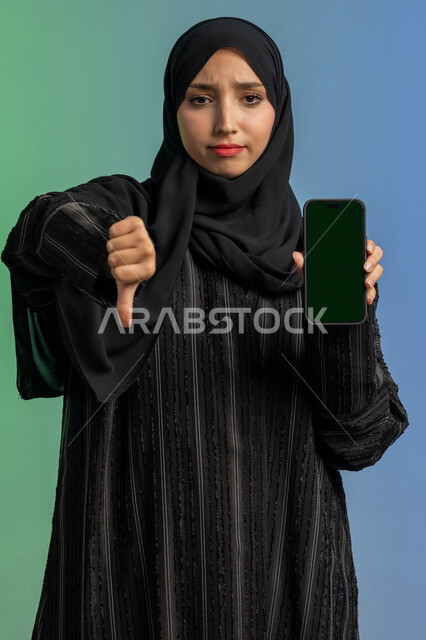 Thumbs down gesture of rejection and disapproval, concept of negative evaluation and annoyance, empty green screen display chroma key on mobile, portrait of veiled Arab Gulf Emirati woman wearing black abaya holding mobile phone in hand, using modern and advanced technical device, colorful background