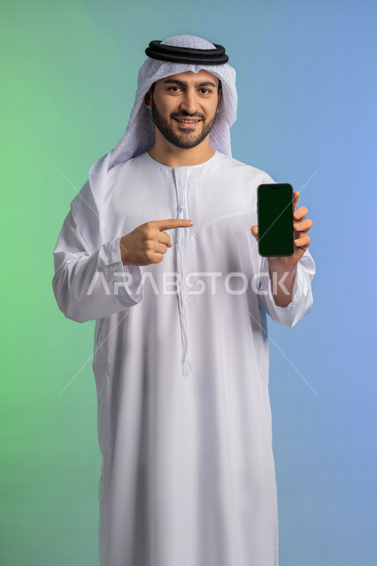 عرض تطبيقات الجوال، الاشارة باصبع السبابة الى شاشة خضراء فارغة كروما،  بورتريه لرجل عربي خليجي اماراتي يرتدي الكندورة والغترة البيضاء يمسك بيده الهاتف المحمول، استخدام جهاز تقني حديث ومتطور،  النظر الى الكاميرا بايماءات السعادة والسرور، خلفية ملونة