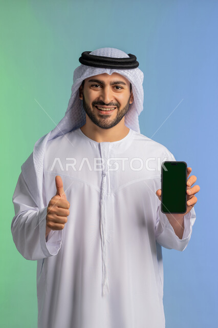 رفع اصبع الابهام للأعلى بايماءات الجودة والامتياز، مفهوم القبول والإعجاب والرضا, عرض تطبيقات الجوال عبر شاشة خضراء فارغة كروما،  بورتريه لرجل عربي خليجي اماراتي يرتدي الكندورة والغترة البيضاء يمسك بيده الهاتف المحمول، استخدام جهاز تقني حديث ومتطور، خلفية ملونة