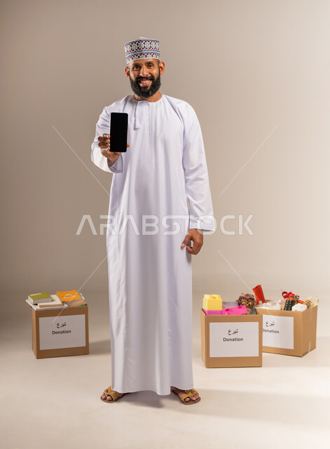 عرض شاشة سوداء فارغة، توفير المستلزمات الضرورية للمحتاجين، مفهوم المساعدة والتبرع، بورتريه لرجل عربي خليجي عماني يرتدي الدشداشة والكمة يقف أمام صناديق التبرعات يمسك الهاتف المحمول ينظر إلى الكاميرا، استخدام جهاز تقني حديث، الجسم بالطول الكامل، خلفية بيج