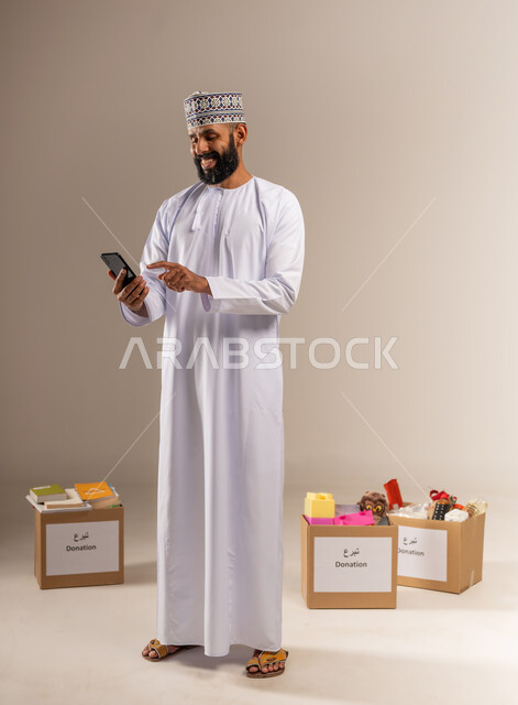 Using a modern technical device, providing essential supplies to the poor and needy, a humanitarian initiative in the Sultanate of Oman, the concept of assistance and donation, portrait of a smiling Omani Gulf Arab man wearing a dishdasha and a cloak standing in front of donation boxes using a mobile phone, full body, beige background