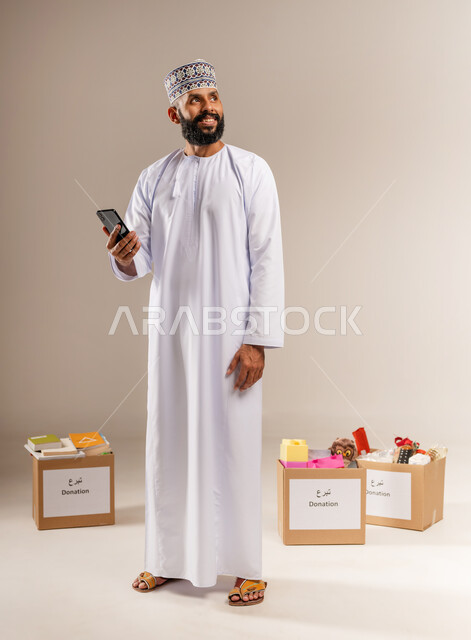 توفير المستلزمات الضرورية للمحتاجين، مبادرة انسانية في سلطنة عمان، مفهوم المساعدة والتبرع، بورتريه لرجل عربي خليجي عماني يرتدي الدشداشة والكمة يقف أمام صناديق التبرعات يمسك الهاتف المحمول ينظر إلى الأعلى، استخدام جهاز تقني حديث، الجسم بالطول الكامل، خلفية بيج