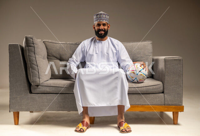 الاستمتاع بمشاهدة الفريق المفضل، قضاء وقت ممتع بمتابعة المباراة، بورتريه لرجل عربي خليجي عماني يرتدي الدشداشة والكمة يجلس على أريكة مريحة معه كرة القدم ينظر إلى الكاميرا بإيماءات السرور والثقة بالنفس، المباريات والألعاب الرياضية، خلفية بيج