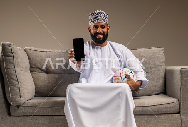 عرض شاشة سوداء فارغة، استخدام جهاز تقني حديث، قضاء وقت ممتع بمتابعة المباراة، بورتريه لرجل عربي خليجي عماني يرتدي الدشداشة والكمة يجلس على أريكة مريحة يمسك كرة القدم والهاتف المحمول ينظر إلى الكاميرا بإيماءات السرور، المباريات والألعاب الرياضية، خلفية بيج