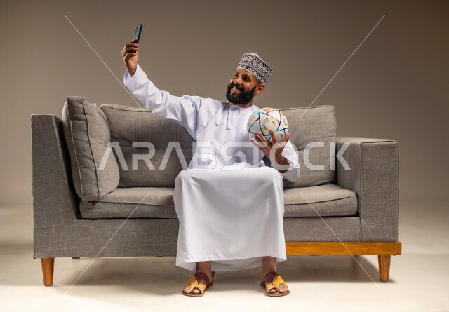 التقاط الصور الذكارية، استخدام جهاز تقني حديث، قضاء وقت ممتع بمتابعة المباراة، بورتريه لرجل عربي خليجي عماني يرتدي الدشداشة والكمة يجلس على أريكة مريحة يمسك كرة القدم والهاتف المحمول يلتقط صورة سيلفي، المباريات والألعاب الرياضية، خلفية بيج