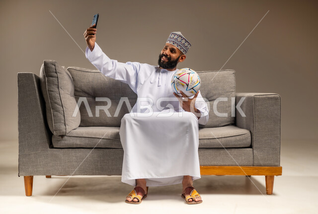 المباريات والألعاب الرياضية، التقاط الصور الذكارية، استخدام جهاز تقني حديث، قضاء وقت ممتع بمتابعة المباراة، بورتريه لرجل عربي خليجي عماني يرتدي الدشداشة والكمة يجلس على أريكة مريحة يمسك كرة القدم والهاتف المحمول يلتقط صورة سيلفي، خلفية بيج