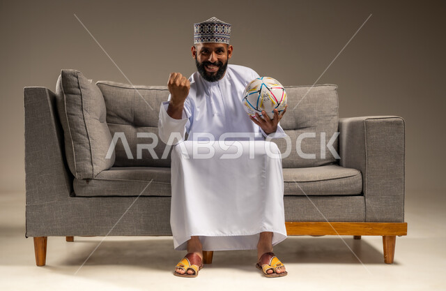 الاستمتاع بمشاهدة الفريق المفضل، قضاء وقت ممتع بمتابعة المباراة، بورتريه لرجل عربي خليجي عماني يرتدي الدشداشة والكمة يجلس على أريكة مريحة يمسك كرة القدم ينظر إلى الكاميرا بتعابير الحماس، رفع قبضة اليد بإيماءات الفوز والانتصار، المباريات والألعاب الرياضية، خلفية بيج