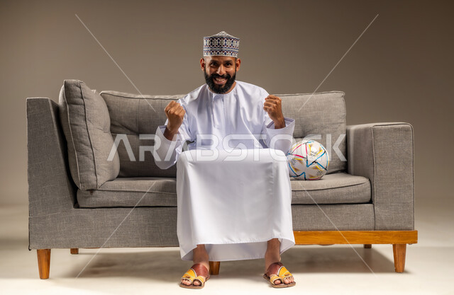 قضاء وقت ممتع بمتابعة المباراة، بورتريه لرجل عربي خليجي عماني يرتدي الدشداشة والكمة يجلس على أريكة مريحة معه كرة القدم ينظر إلى الكاميرا بتعابير الحماس، الاستمتاع بمشاهدة الفريق المفضل، رفع قبضتي اليدين بإيماءات الفوز والانتصار، المباريات والألعاب الرياضية، خلفية بيج